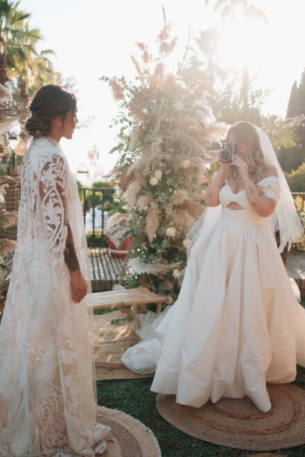 La novia del velo fotografía a la otra con una cámara instantánea junto a un arreglo de pampas