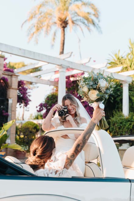 La novia del velo fotografía desde el descapotable a la otra, que levanta el ramo