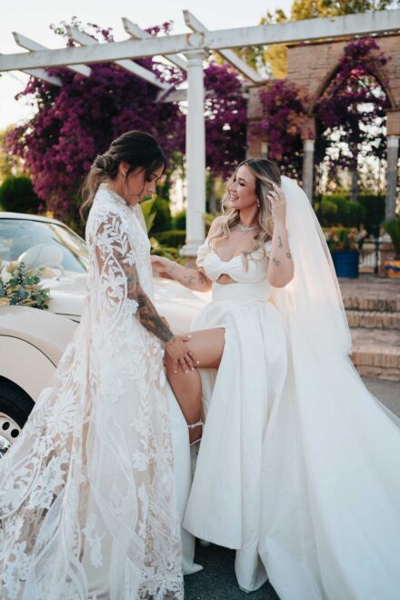 La novia del velo asoma la pierna junto al descapotable y la otra le apoya la mano en la rodilla, bajo las buganvillas