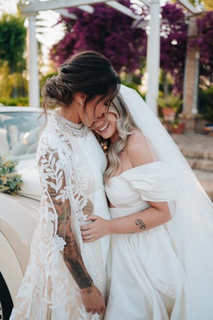 Las novias se abrazan de cerca y una esconde la risa en el cuello de la otra, junto al descapotable