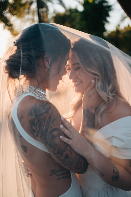 Las novias juntan las frentes bajo el velo a contraluz, una con la mano en el hombro tatuado de la otra