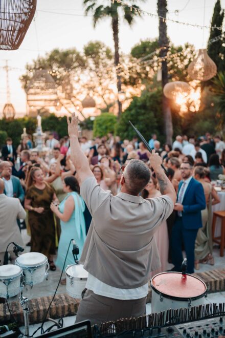 Un percusionista de espaldas alza los brazos con una baqueta frente a los invitados que bailan al atardecer