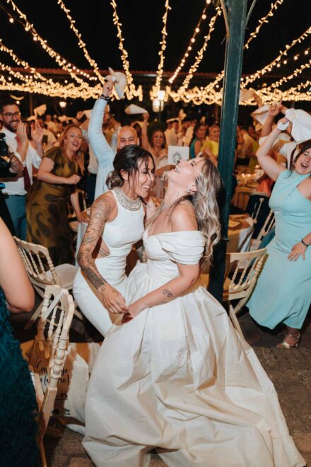 Una de las novias echa la cabeza atrás riendo mientras baila con la otra entre servilletas al aire