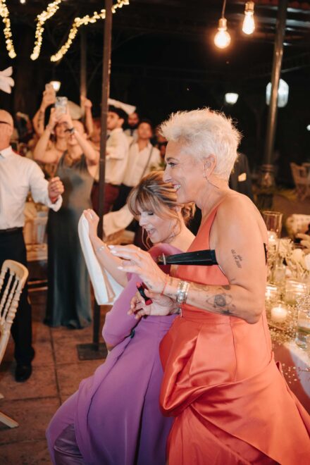 Dos invitadas, una de vestido coral y otra de lila, bailan sonrientes junto a las mesas del banquete