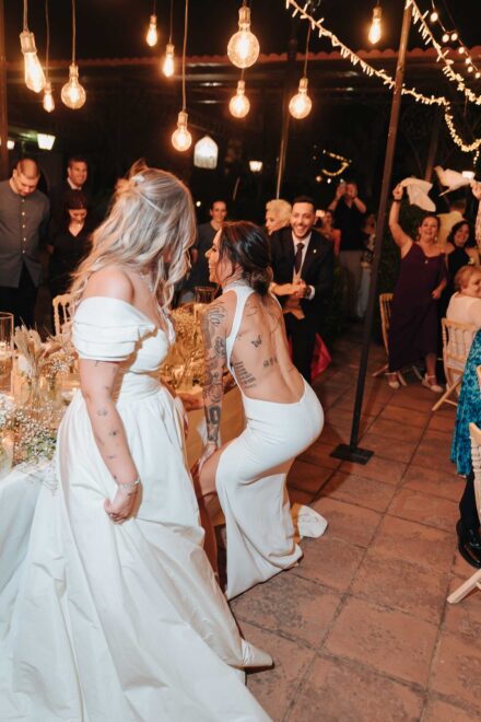 Una novia con la espalda tatuada baila agachándose frente a la otra junto a la mesa, bajo bombillas colgantes