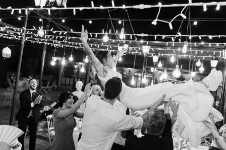 Los invitados mantean a una de las novias bajo las guirnaldas de luces, en blanco y negro