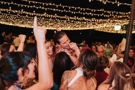 Una invitada canta con un micrófono rodeada de invitados que levantan los brazos, con una novia de espaldas