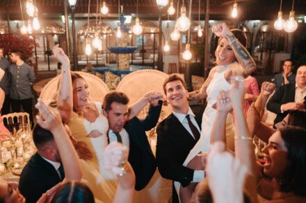 Un invitado levanta a una de las novias, que alza el brazo tatuado, ante los sillones de mimbre y la fuente del patio