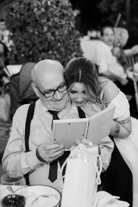 Una de las novias abraza emocionada a un invitado mayor que mira un marco de regalo, en blanco y negro