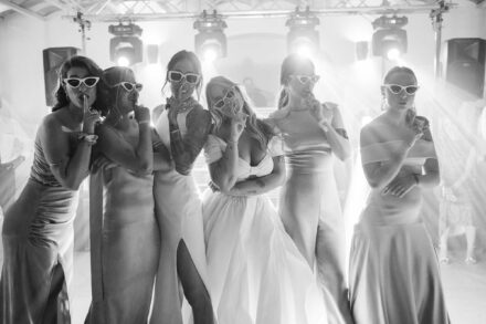 Las novias y cuatro invitadas con gafas de sol blancas se llevan el dedo a los labios, en blanco y negro