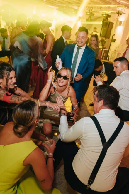 Andrea Medina, con gafas de sol oscuras, baila sentada en el suelo en un corro de invitados