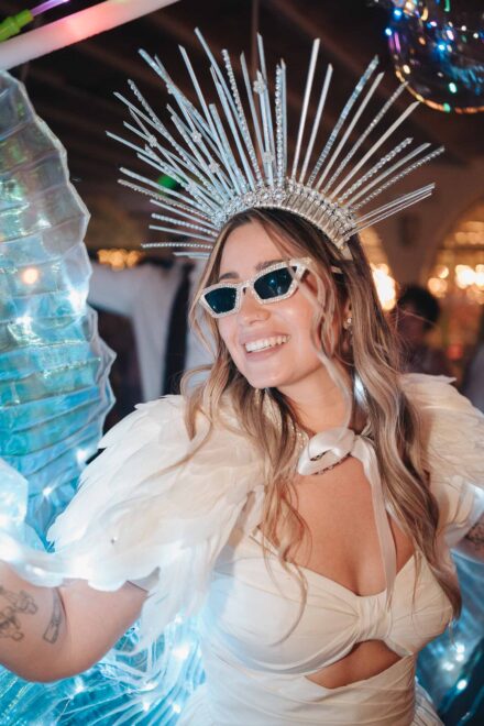 Una de las novias sonríe de cerca con corona de pinchos plateados, gafas de sol brillantes y alas de plumas
