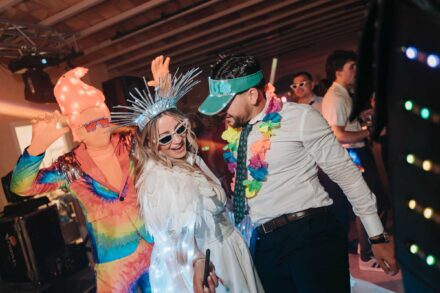 Una novia con corona plateada baila con un invitado con collar de flores y alguien disfrazado de arcoíris