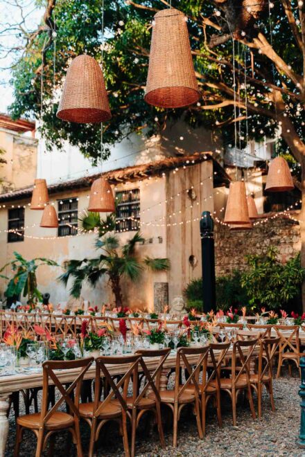 Mesas largas en un patio de grava bajo lámparas de mimbre colgadas de un árbol, con flores rojas