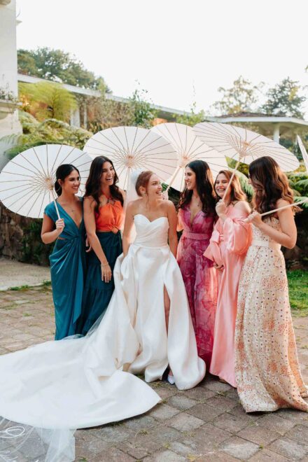 La novia se ríe rodeada de cinco invitadas con vestidos de colores y sombrillas de papel blancas