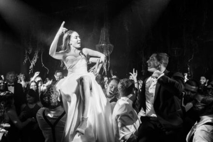 La novia, subida en una silla a hombros de los invitados, baila con el dedo en alto, en blanco y negro