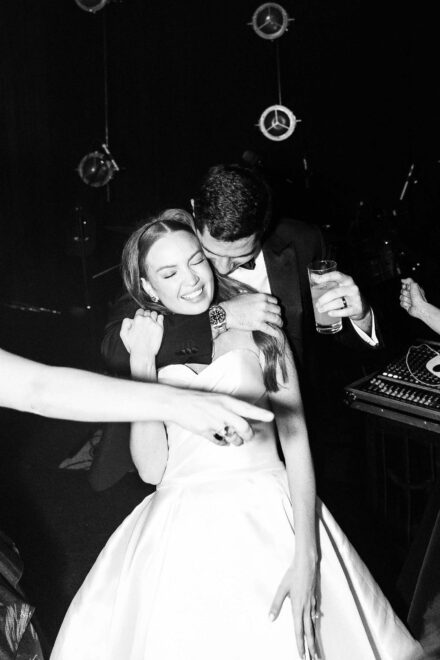 El novio abraza por detrás a la novia, que sonríe con los ojos cerrados junto a la mesa de mezclas, en blanco y negro