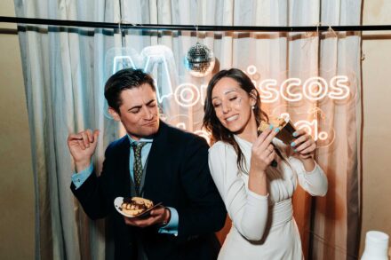 Los novios bailan con un dulce en la mano delante de una cortina con letrero de neón y bola de espejos