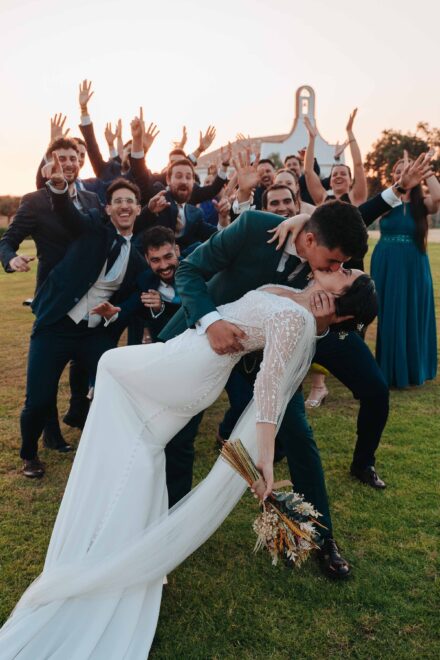 El novio besa a la novia inclinándola hacia atrás en el césped mientras los invitados levantan las manos, al atardecer