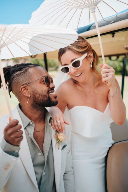 Los novios, con gafas de sol, bajo sombrillas de papel blancas en un carrito de golf; ella se ríe mirando abajo