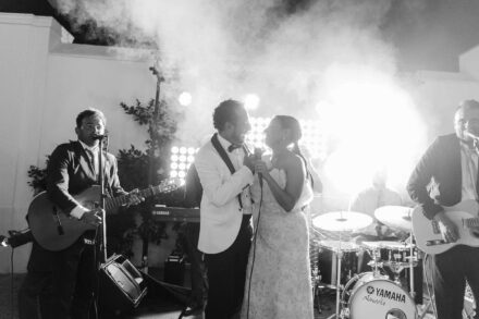 Los novios cantan juntos al micrófono en el escenario con la banda, entre humo, en blanco y negro