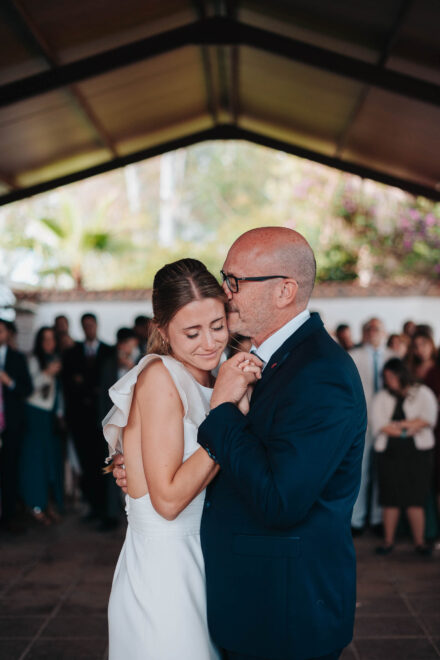 La novia, con los ojos cerrados, baila abrazada a un hombre con gafas que le besa la cabeza bajo un porche