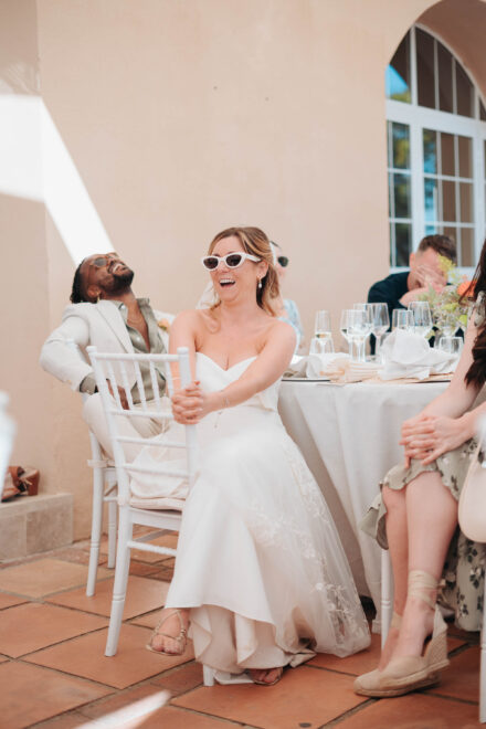 La novia, con gafas de sol blancas, se ríe sentada a la mesa del banquete mientras un invitado echa la cabeza atrás