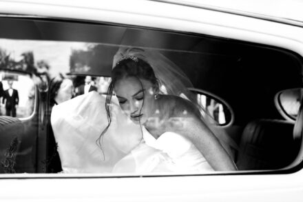 La novia, con tiara y velo, mira hacia abajo dentro de un coche antiguo, tras la ventanilla, en blanco y negro