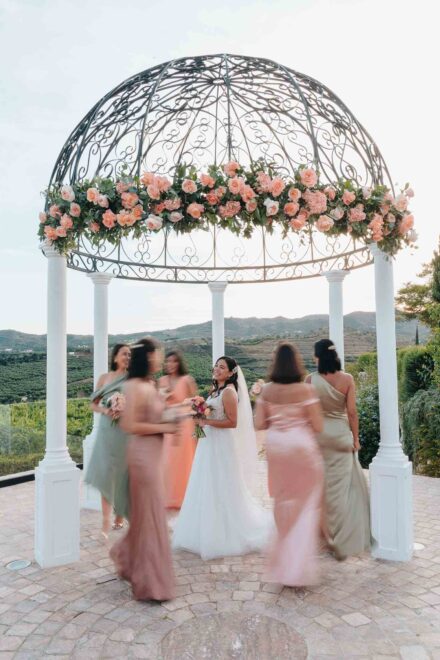 La novia ríe bajo un templete de forja con rosas mientras amigas con vestidos pastel giran a su alrededor