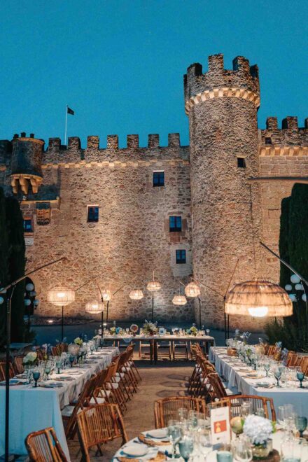 Mesas del banquete con lámparas de mimbre frente a un castillo de piedra almenado e iluminado, al anochecer