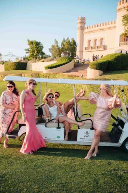 La novia y sus amigas se ríen alrededor de un carrito de golf en el césped, al pie de un edificio almenado con torres