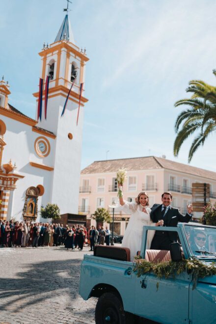 Los novios saludan desde un todoterreno descapotable azul frente a la iglesia, con los invitados en la plaza