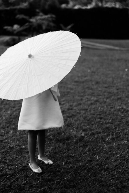 Una niña con vestido blanco se tapa con una sombrilla de papel sobre el césped, en blanco y negro