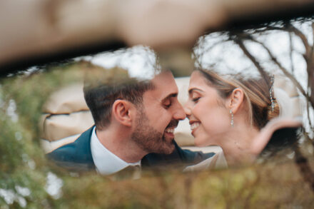 Los novios sonríen nariz con nariz, vistos entre ramas desenfocadas, con la capota plegada del coche detrás