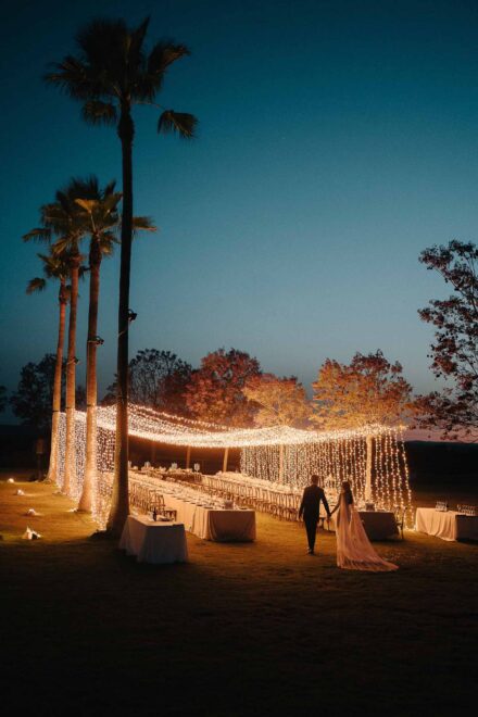 Los novios caminan de noche hacia las mesas del banquete, cubiertas por cortinas de luces entre palmeras