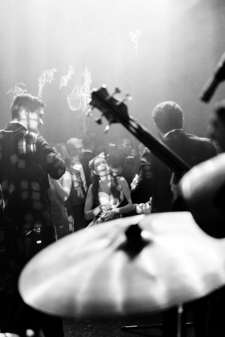 La novia ríe con los ojos cerrados entre los invitados, vista tras el platillo y el bajo, en blanco y negro