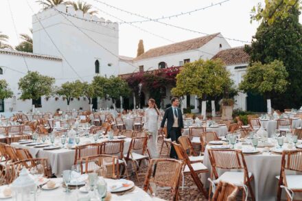 Los novios pasean entre las mesas del banquete en un patio blanco con buganvillas y guirnaldas de luces