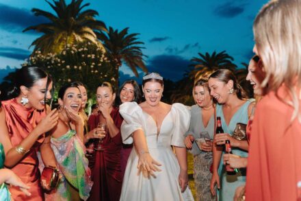 La novia, con tiara y mangas abullonadas, se ríe rodeada de amigas con copas y cervezas al anochecer entre palmeras