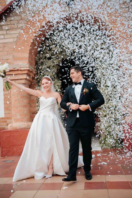 Los novios salen bajo una nube de confeti blanco por un arco de ladrillo, ella con el ramo en alto