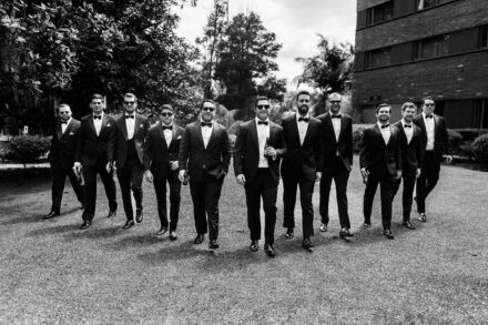 Un grupo de hombres con esmoquin y pajarita, varios con gafas de sol, camina por el césped, en blanco y negro