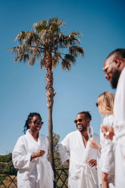 Amigos en albornoz blanco y gafas de sol se ríen con sus copas bajo una palmera, en los preparativos