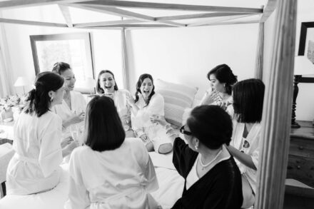 Mujeres en bata ríen y brindan sentadas en una cama con dosel durante los preparativos, en blanco y negro