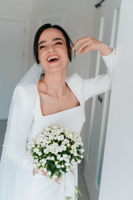 La novia se ríe a carcajadas apoyada en la pared, con velo de perlas y un ramo de flores blancas