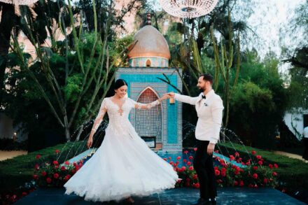 La novia gira el vestido de la mano del novio, de chaqueta blanca, ante un templete de azulejos turquesa y surtidores