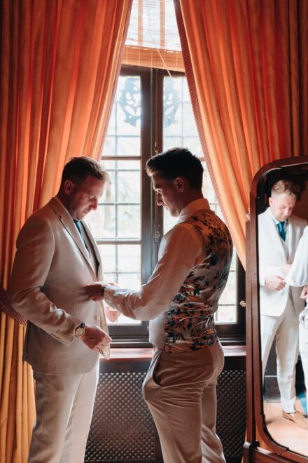 Groom getting ready with help adjusting his beige suit by the window at Hacienda Villa Luisa in Seville