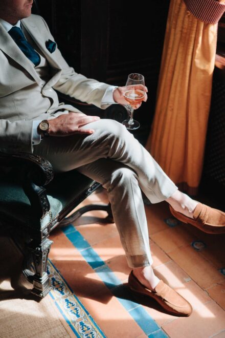 Groom sitting by the window holding a glass, beige suit details at Hacienda Villa Luisa wedding Seville