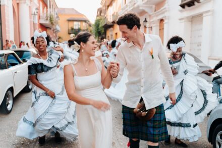 Los novios, él con kilt, avanzan de la mano por una calle colonial entre bailarinas de blanco con volantes