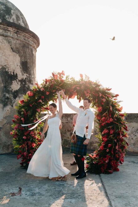 Los novios giran de la mano bajo un arco de flores rojas, junto a una garita de piedra, con un pájaro al vuelo