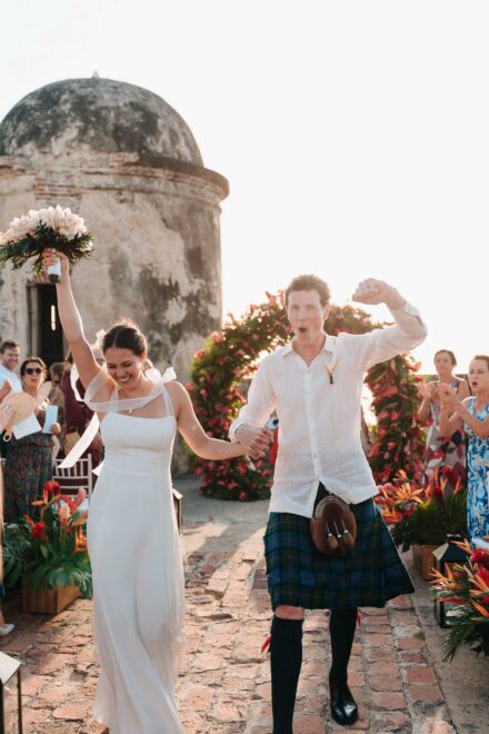 Los novios, él con kilt, alzan ramo y puño junto a una garita de piedra y un arco de flores rojas