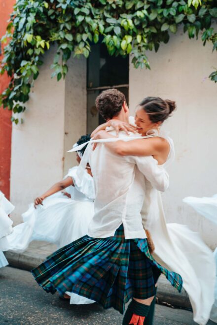 Los novios se abrazan girando en la calle, con el kilt de él y las faldas blancas de las bailarinas al vuelo